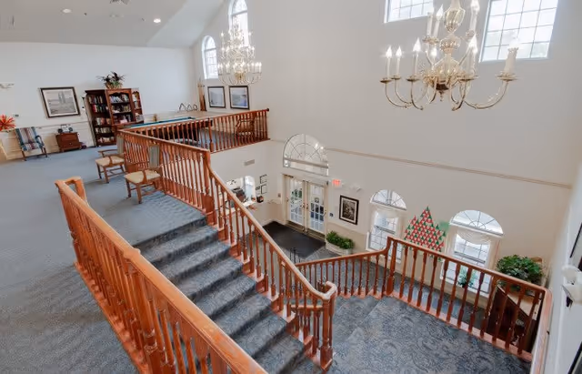 Interior view of a senior living facility featuring a carpeted staircase with wooden railings leading down to a lobby area. The space is well-lit with large arched windows and elegant chandeliers. There is a seating area with chairs and a bookshelf on the upper level, and decorative plants and framed pictures adorn the walls.