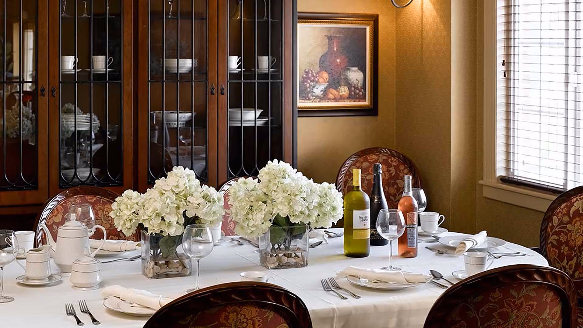 A formal dining room with a table set for a meal, featuring white tablecloth, white plates, silverware, glassware, and napkins. The table is decorated with three vases of white flowers and three bottles of wine. In the background, there is a wooden china cabinet with glass doors displaying dishes and cups, and a framed painting on the wall. A window with blinds allows natural light into the room.