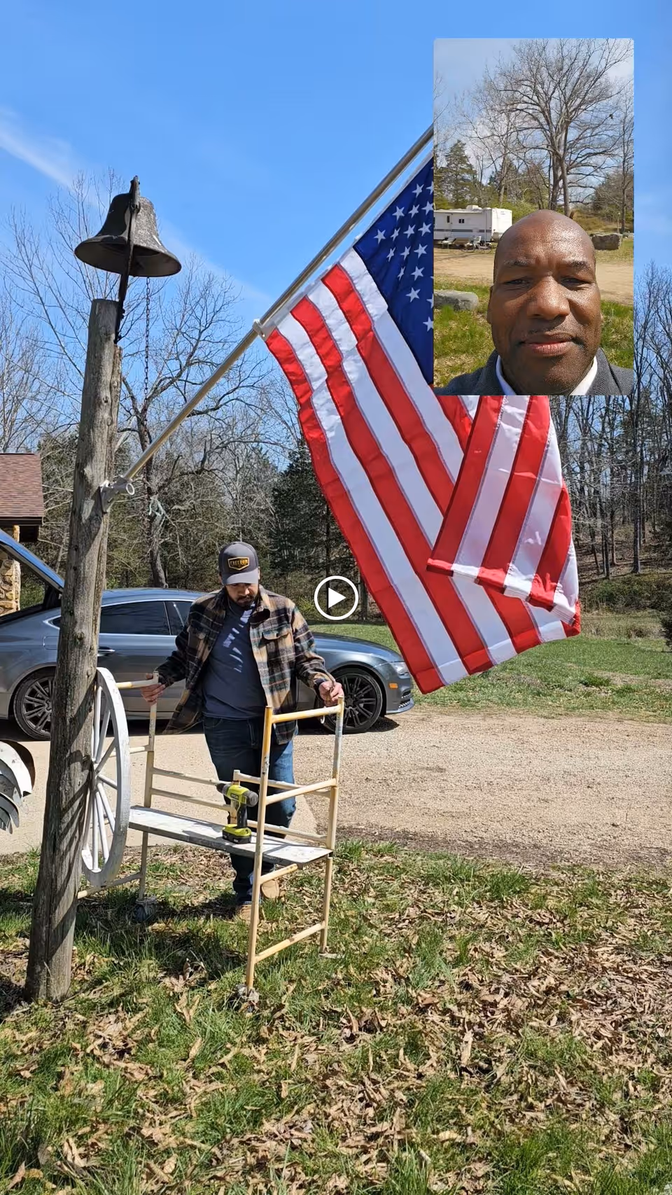 An outdoor scene showing a man by a wagon wheel and small scaffold under a large American flag on a pole, with a parked car and trees in the background and a small inset selfie of another man in the top-right corner.