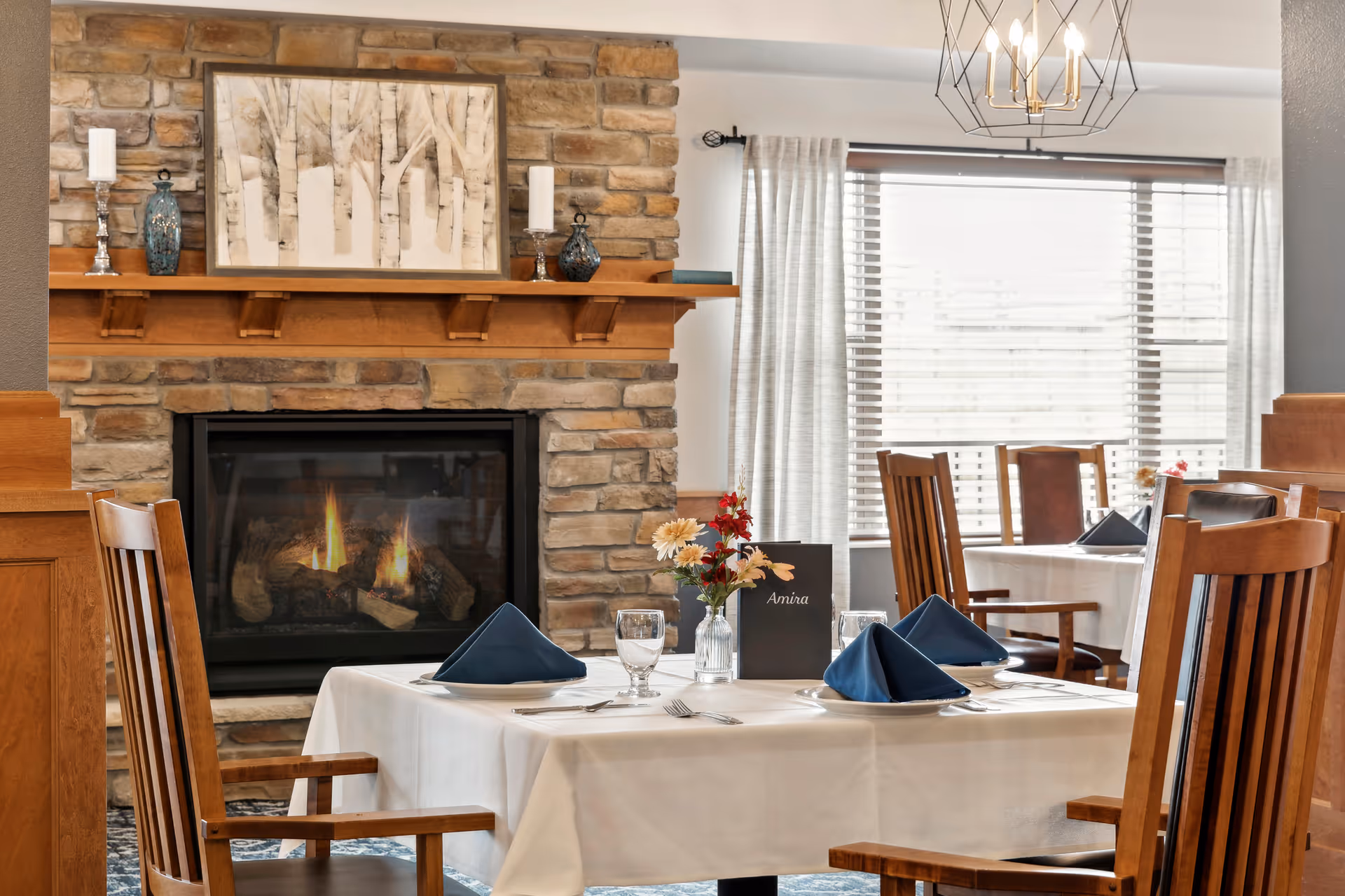 A cozy dining area with wooden chairs and tables covered with white tablecloths, set with blue folded napkins, glassware, and silverware. A stone fireplace with a lit fire and a wooden mantel decorated with candles and vases is visible in the background. Large windows with blinds and light curtains allow natural light into the room.