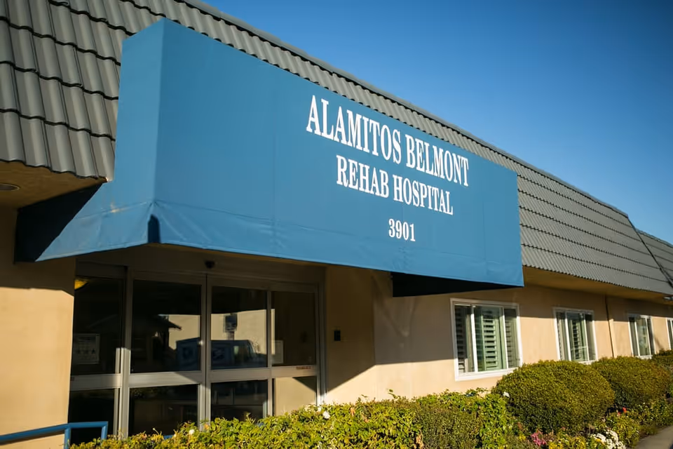 Exterior view of Alamitos Belmont Rehab Hospital showing the entrance with a blue awning displaying the facility name and address number 3901. The building has beige walls, a tiled roof, and bushes along the front.