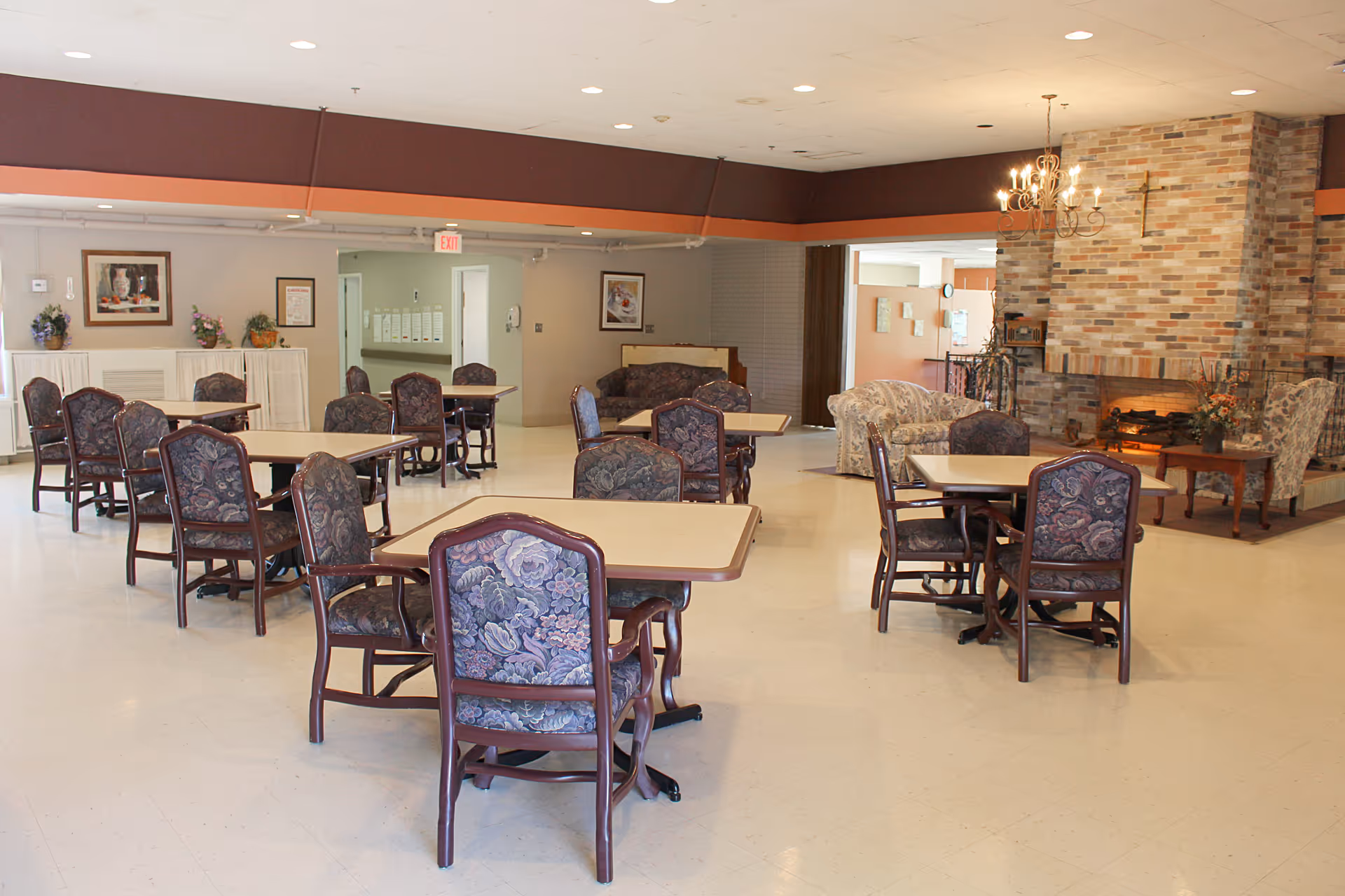 Spacious senior living common room with multiple dining tables and upholstered chairs and a brick fireplace seating area.