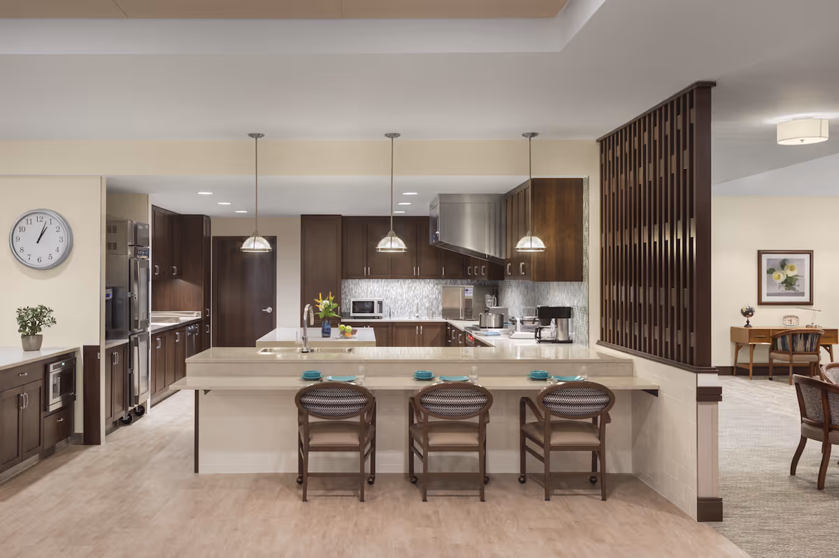 Spacious communal kitchen with a central island, three bar stools, pendant lights, stainless-steel appliances, and dark wood cabinets.