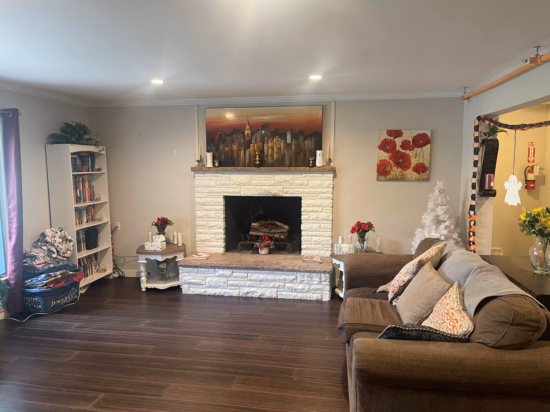 A cozy living room with a brown sofa adorned with patterned cushions, a white brick fireplace with a cityscape painting above it, two side tables with flowers and candles, a white bookshelf filled with books, and a small white decorative tree. The room has wooden flooring and neutral-colored walls.
