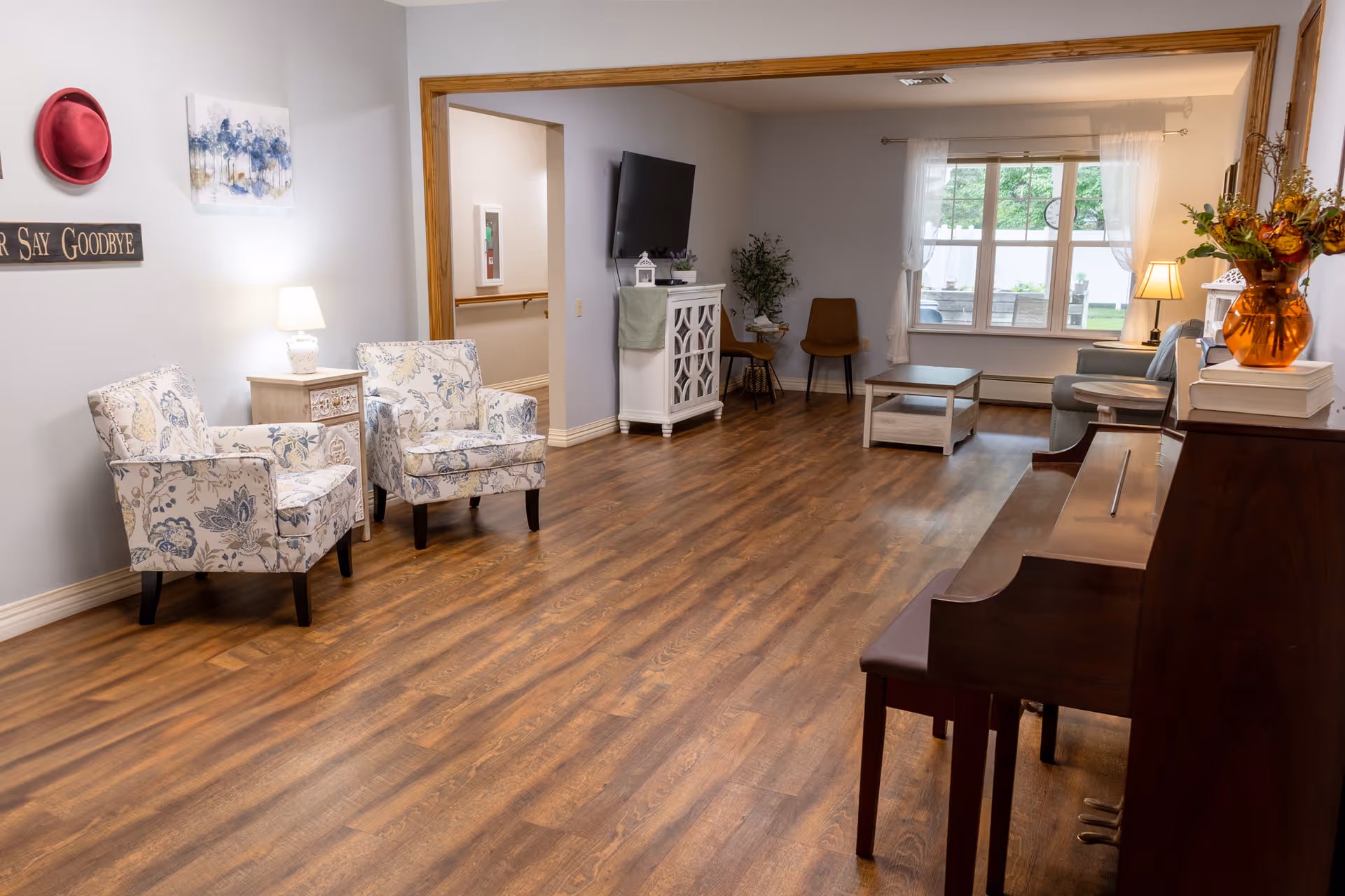 A cozy senior living common area with wooden flooring, two floral armchairs next to a small side table with a lamp, a wall-mounted TV, a white cabinet, two brown chairs, a coffee table, a gray sofa near a window with white curtains, and a wooden piano with a vase of flowers on top.