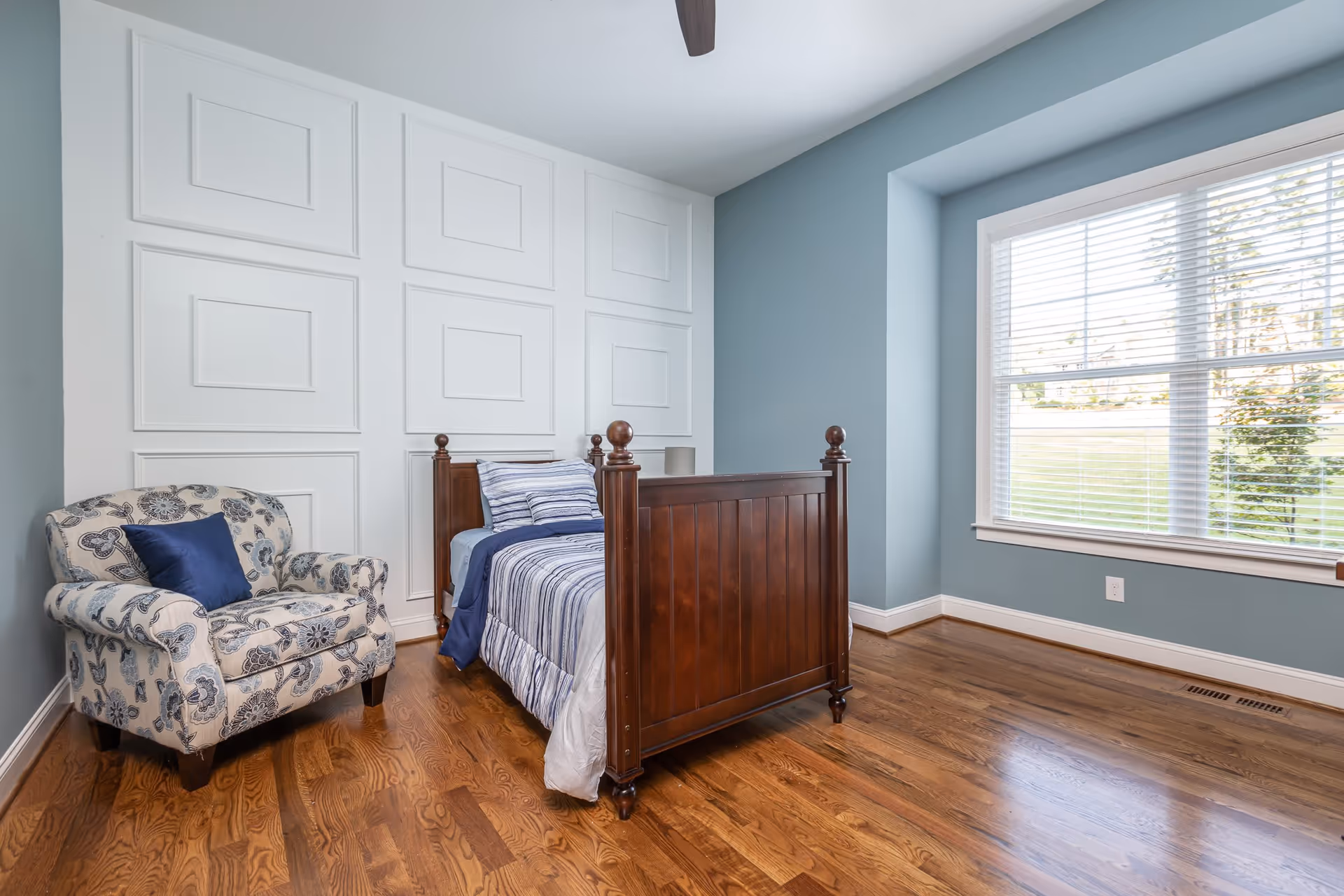 A bedroom with a wooden single bed dressed in blue and white striped bedding, a patterned armchair with a blue pillow, light blue walls, white decorative wall panels, a large window with white blinds, and hardwood flooring.