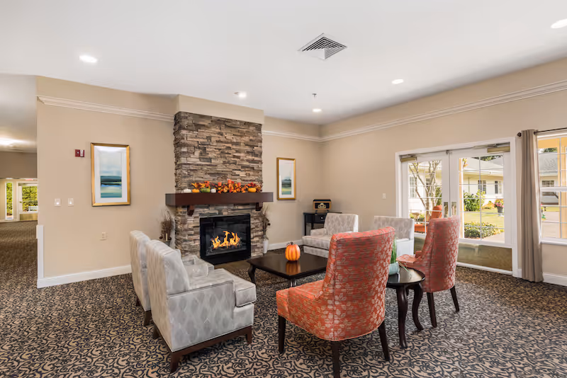 A cozy living room area in Inland Point Retirement Community featuring a stone fireplace with a mantel decorated with autumn-themed items. The room has a patterned carpet and is furnished with two red patterned armchairs, four light gray armchairs, and two dark wood coffee tables. Two framed landscape paintings hang on the walls, and large windows and glass doors provide a view of the outdoor garden area.