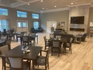 A spacious senior living facility common area with multiple dark wood tables and chairs arranged throughout the room. The room features large windows allowing natural light, a stone fireplace with a mounted flat-screen TV above it, and a piano in the corner. The flooring is light wood, and the walls are painted in neutral tones.