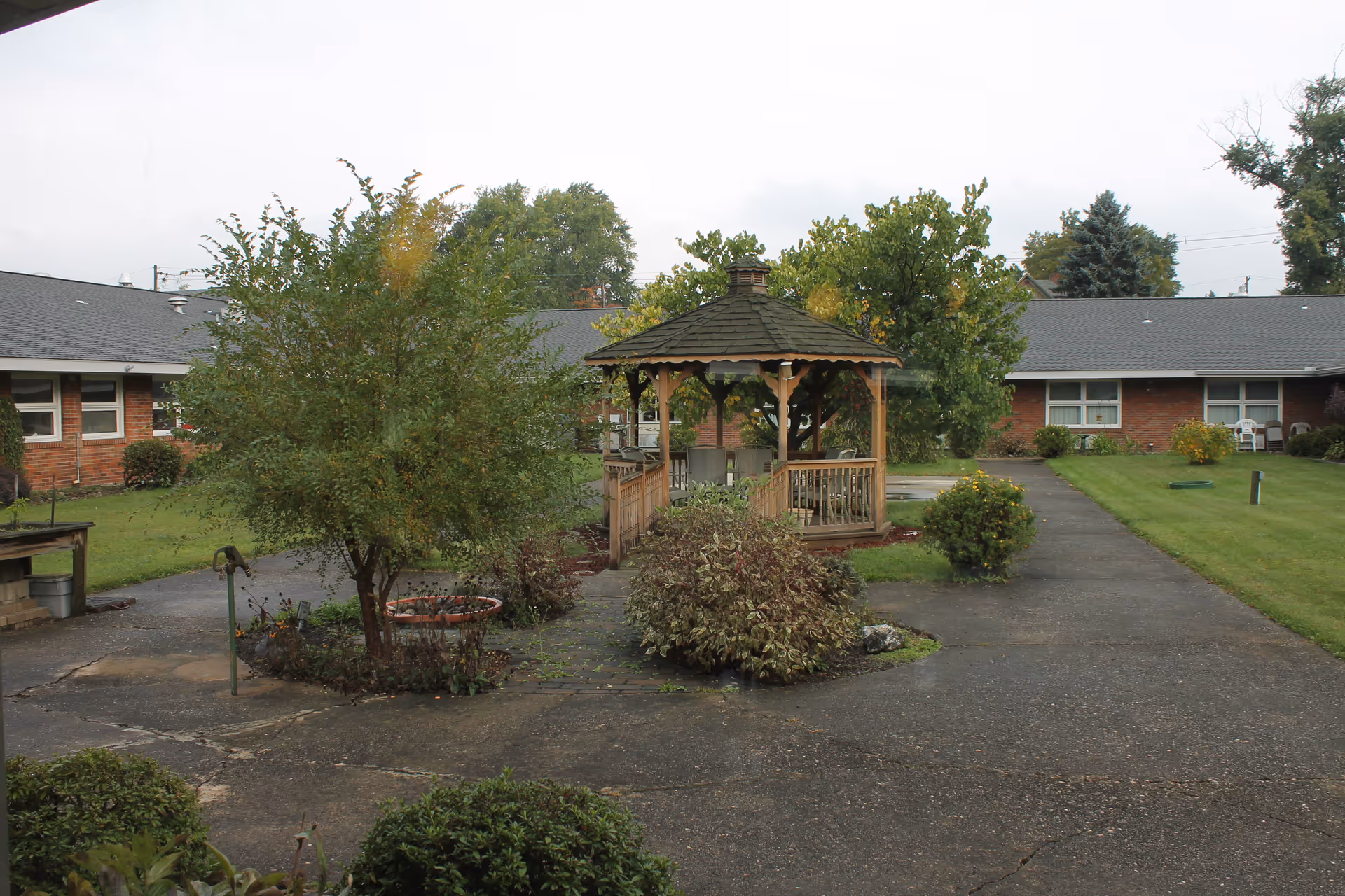 Outdoor courtyard area of a healthcare and rehabilitation center featuring a wooden gazebo surrounded by trees, bushes, and paved walkways. Single-story brick buildings with windows and a gray roof enclose the courtyard.