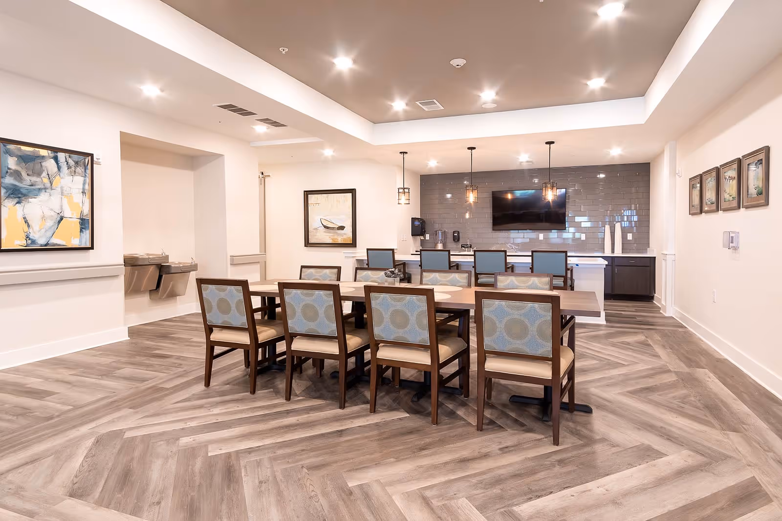 A modern dining room with a long wooden table surrounded by eight chairs with blue patterned backs and beige seats. The room features a light wood herringbone floor, white walls with framed artwork, a wall-mounted TV, and a counter area with pendant lights hanging above.