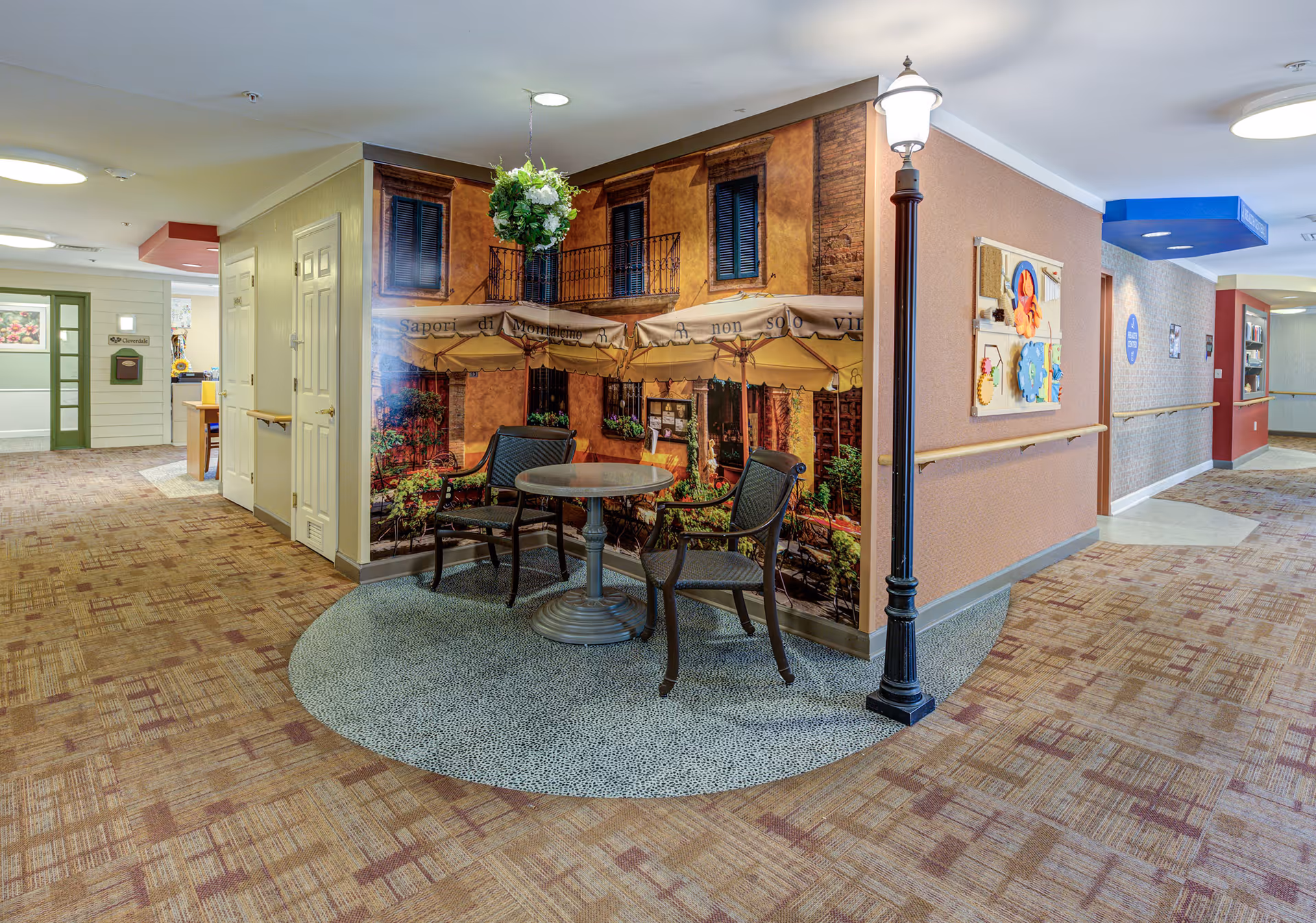 Hallway seating area in a senior living facility with a small round table and chairs set against a mural of an outdoor café.