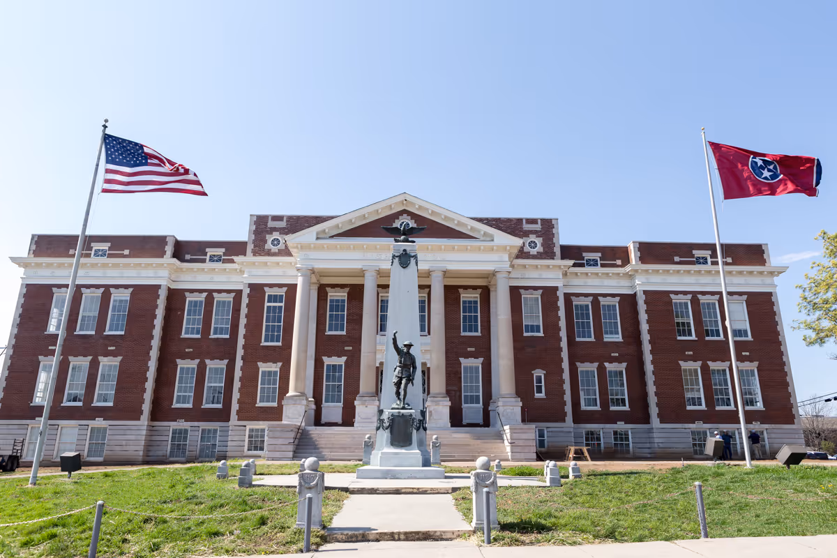 Front exterior view of a large red brick building with white columns and multiple windows, flanked by two flagpoles flying the American flag and the Tennessee state flag. A statue of a soldier stands in the center on a pedestal in front of the building.