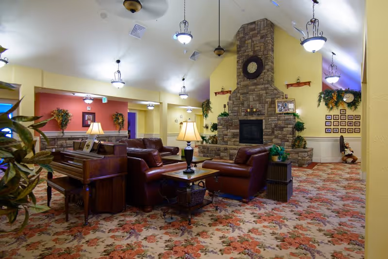 A cozy assisted living facility common area with floral carpet, leather sofas, a wooden piano, and a stone fireplace with a clock above it. The walls are painted yellow and decorated with plants, framed pictures, and wall sconces. Several hanging light fixtures illuminate the room.