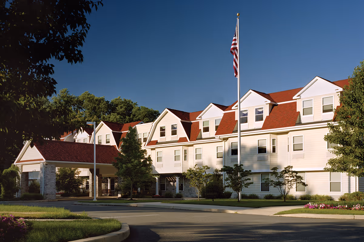 Exterior view of a senior living facility building with white siding and red roof shingles, surrounded by trees and landscaping. An American flag is flying on a flagpole in front of the building under a clear blue sky.