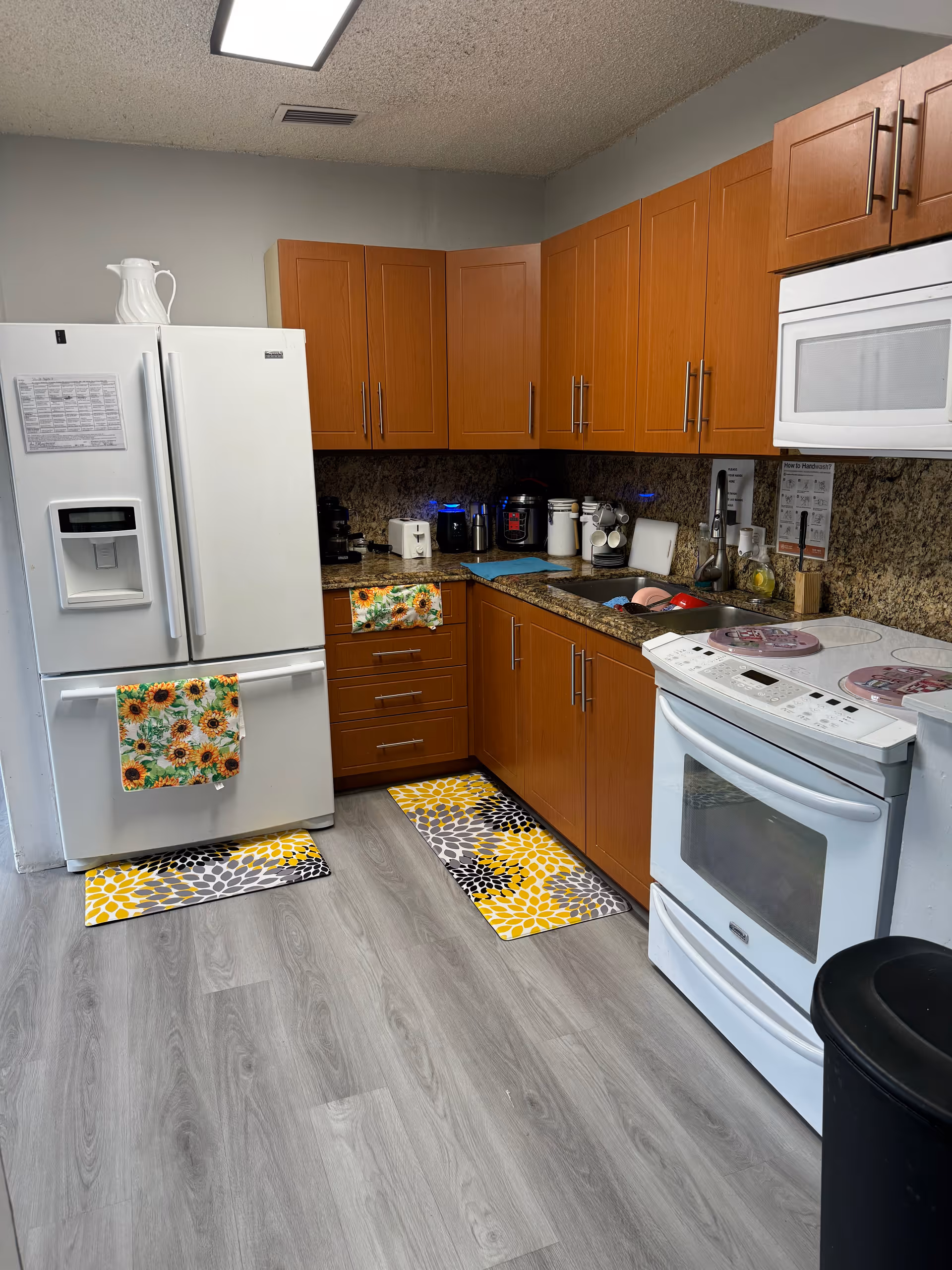 A kitchen with wooden cabinets, a white refrigerator with sunflower-patterned towels, a white stove with pink covers on the burners, a microwave above the stove, and various kitchen appliances on the granite countertop. The floor has light gray wood-like flooring with two colorful rugs featuring yellow, gray, and black patterns.
