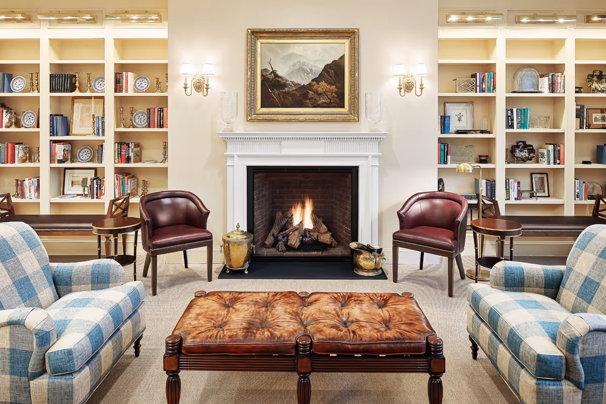 A cozy living room with a lit fireplace in the center, flanked by built-in bookshelves filled with books and decorative items. Two blue and white checkered armchairs face the fireplace, with two brown leather chairs and a tufted leather ottoman in between. Above the fireplace hangs a framed painting of a mountainous landscape. The room is softly lit by wall sconces and overhead lights.