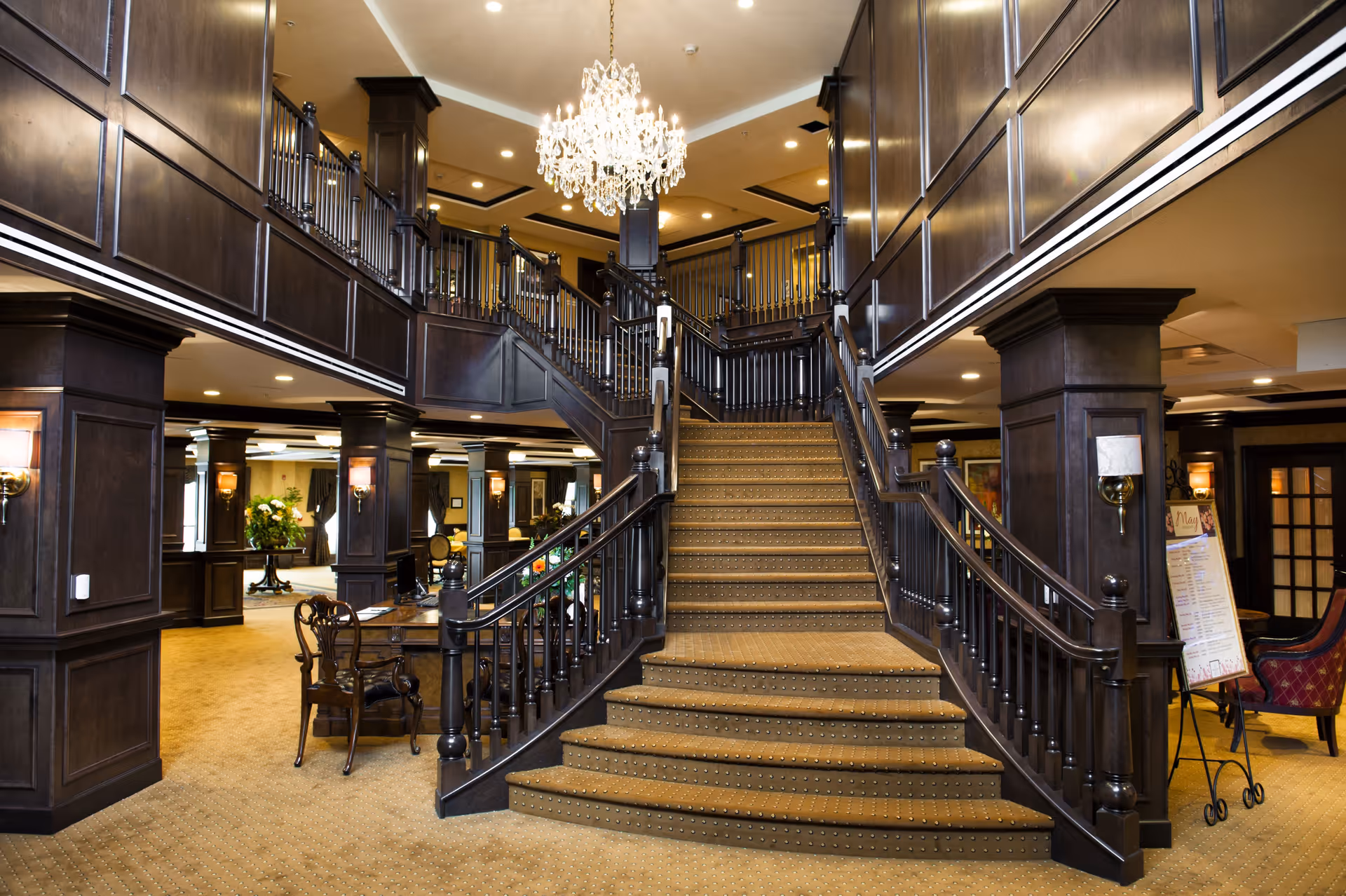 Interior view of a senior living facility featuring a grand staircase with dark wood railings and paneling, a large crystal chandelier hanging from the ceiling, and a carpeted floor. The area includes seating arrangements and decorative lighting fixtures on the walls.