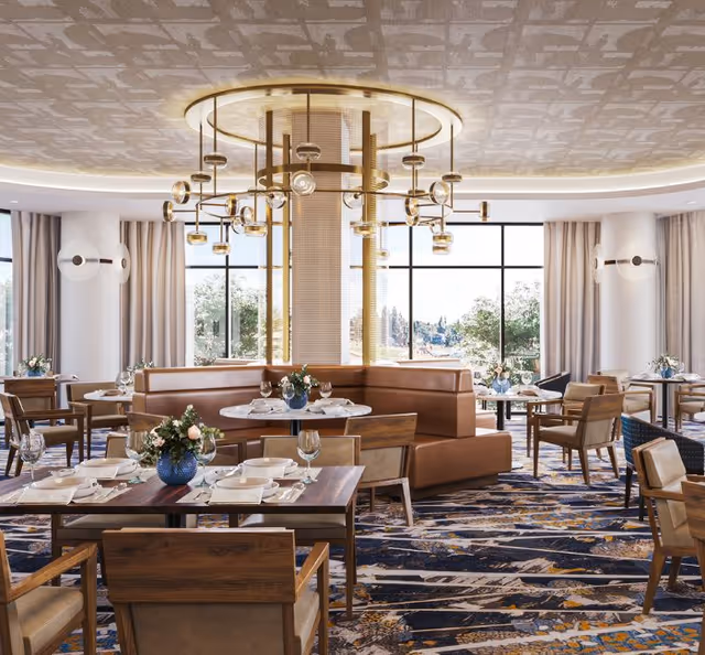 A bright and elegant dining room with large windows letting in natural light. The room features wooden tables set with white plates, napkins, and glassware. There are comfortable wooden chairs and a curved brown leather booth seating around a central column with a modern chandelier above. The carpet has a patterned design in blue, beige, and gold tones, and there are floral centerpieces on the tables.