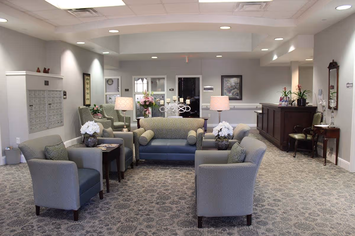 Lobby seating area in a senior living facility with sofas, armchairs, side tables, lamps, flowers, and a reception desk.