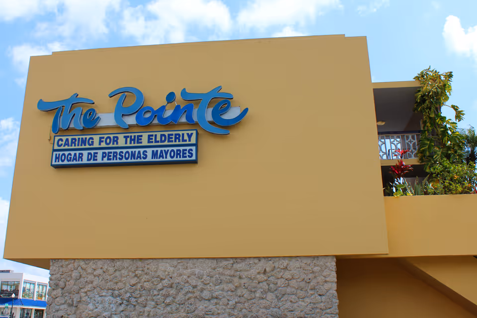 Exterior of a yellow building displaying a blue sign that reads 'The Pointe' with a smaller sign 'Caring for the Elderly / Hogar de Personas Mayores'.