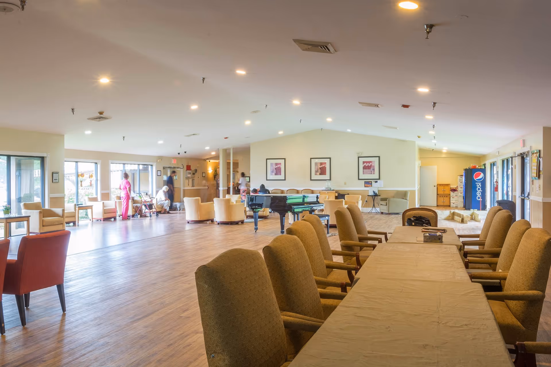 A spacious common area in a senior living facility with multiple seating arrangements including armchairs and a long table with chairs. There is a grand piano in the center, large windows letting in natural light, and several people, including staff and residents, interacting in the background. A Pepsi vending machine is visible on the right side near the exit doors.