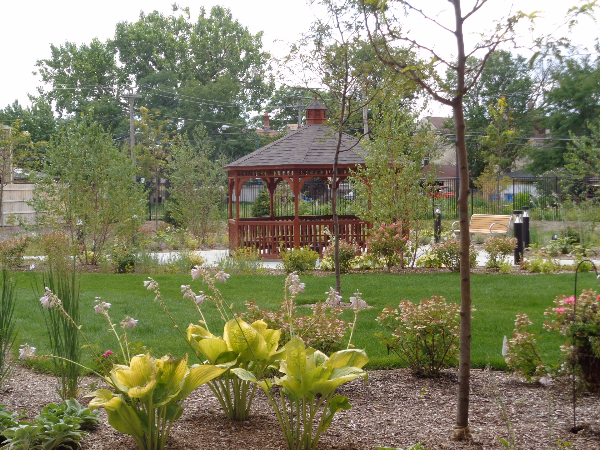 A well-maintained garden area with green grass, various plants, and flowers in the foreground. In the center background, there is a wooden gazebo with a shingled roof. Trees and shrubs surround the area, and a wooden bench is visible near the gazebo. Power lines and residential houses can be seen in the distant background.