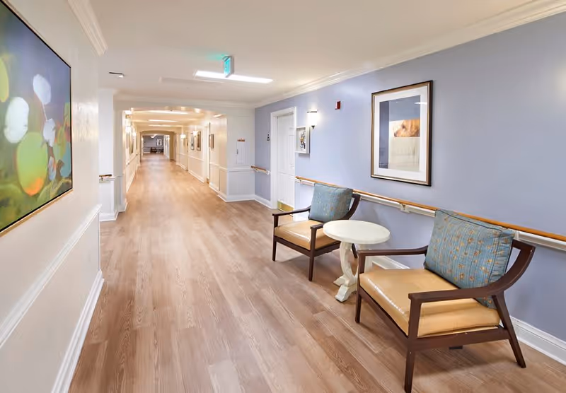 A long, well-lit hallway in a senior living facility with light wood flooring and light blue walls. Two cushioned chairs with blue pillows and a small round white table are placed against the right wall. Handrails run along both sides of the hallway. Several framed pictures hang on the walls, and an exit sign is visible on the ceiling.