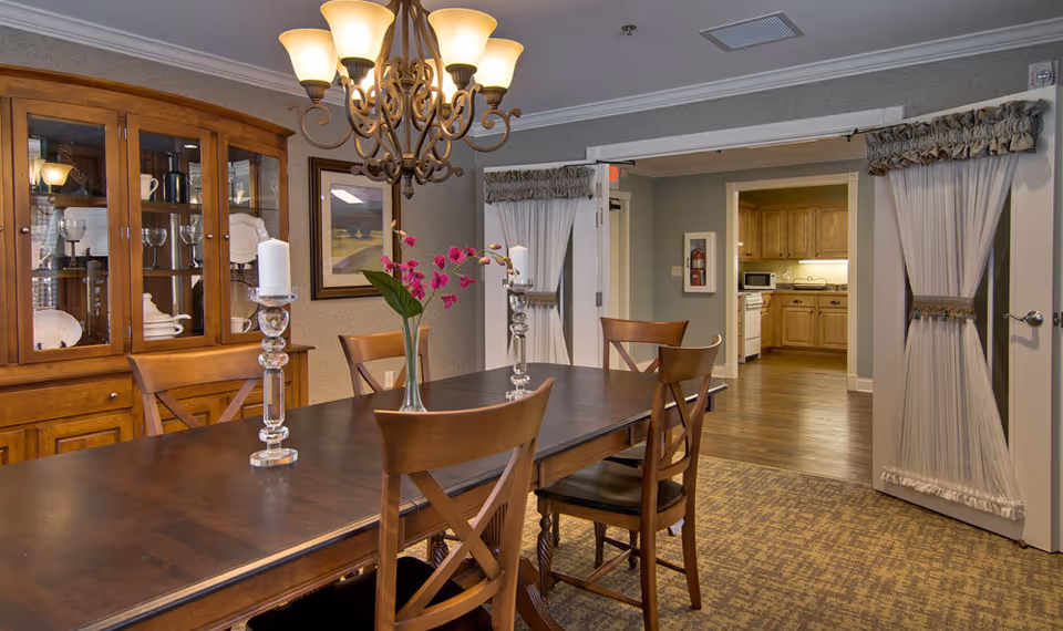 A dining room with a dark wooden table and four wooden chairs. On the table are two glass candle holders with white candles and a vase with pink flowers. A wooden cabinet with glass doors displaying dishes and glassware is against the wall. A chandelier with multiple lights hangs above the table. In the background, an open doorway with white curtains leads to a kitchen area with wooden cabinets and appliances.