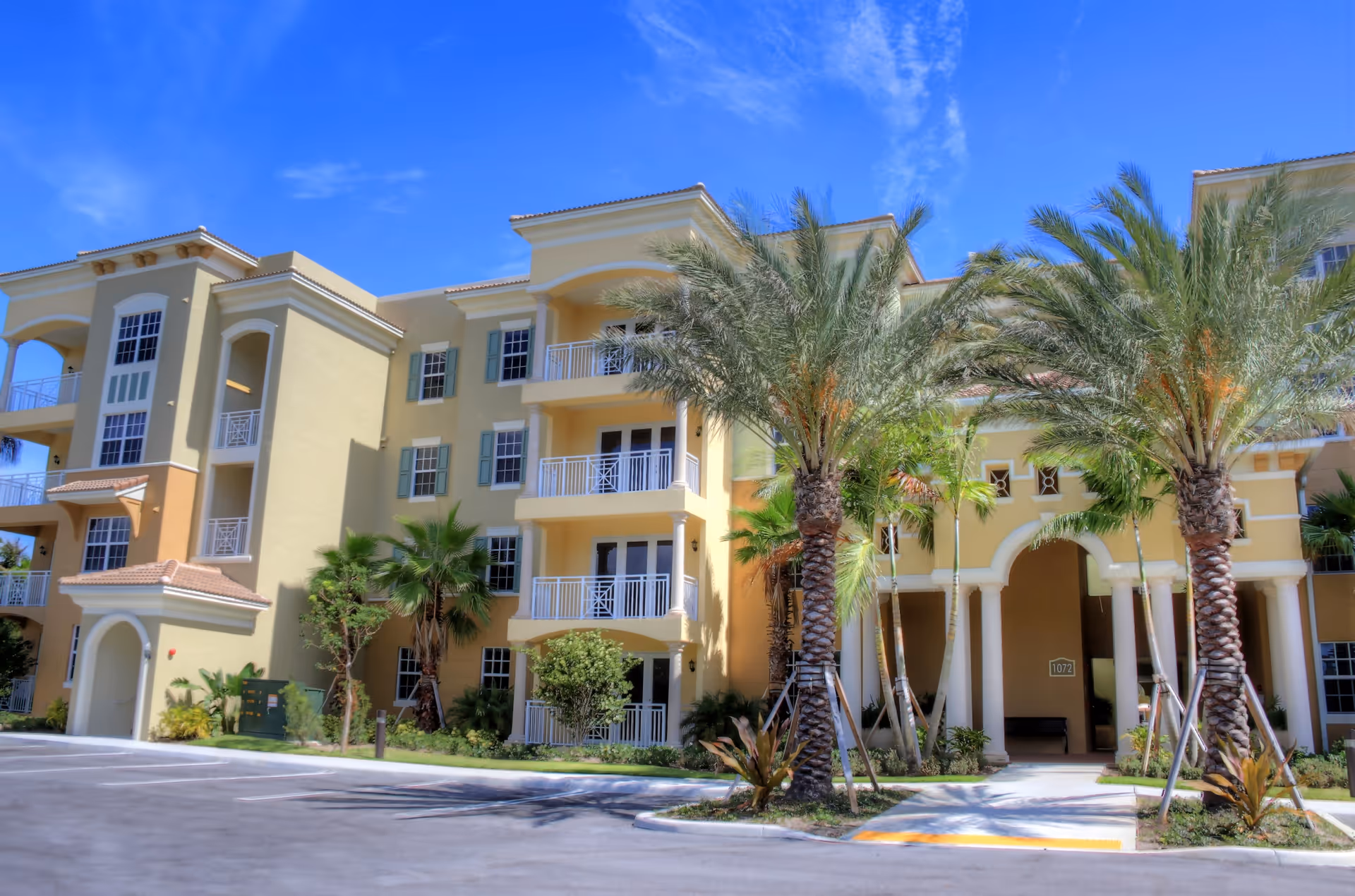 Front view of a multi-story yellow Mediterranean-style building with balconies, palm trees, and an arched entrance under a blue sky.