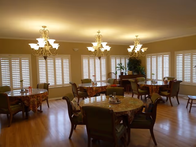 A dining room with several round tables covered with floral tablecloths, each surrounded by green upholstered chairs. The room has wooden flooring, large windows with white plantation shutters, and three ornate chandeliers hanging from the ceiling. There are plants and a piano against the far wall.