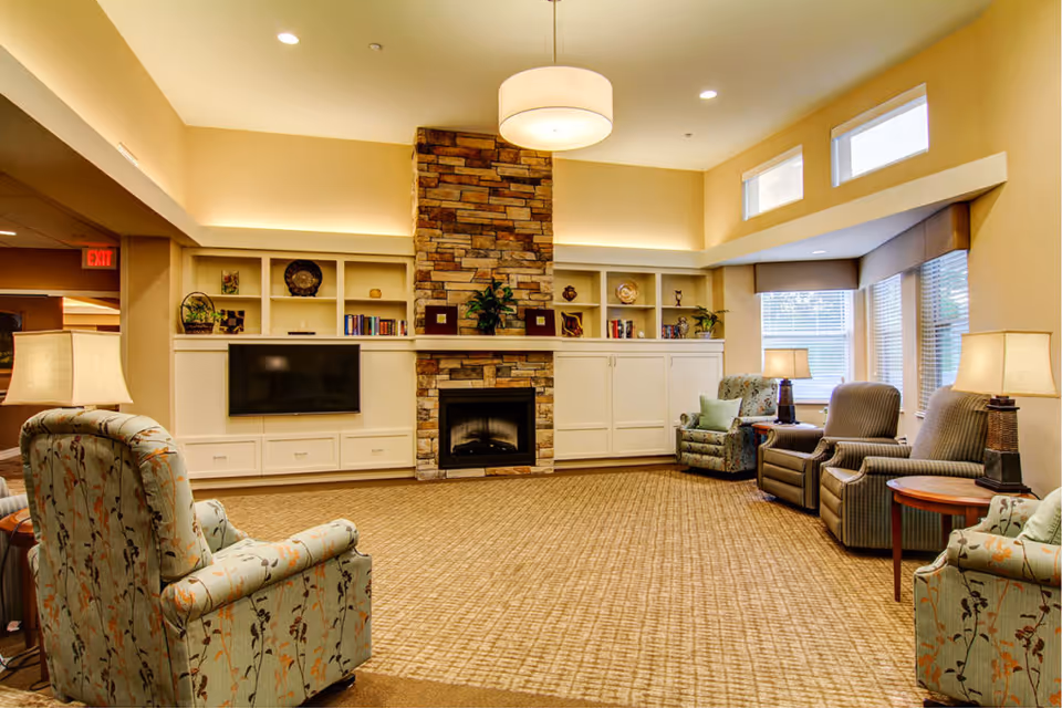 Communal living room with upholstered armchairs arranged around a stone fireplace, built-in shelves, and a wall-mounted TV.