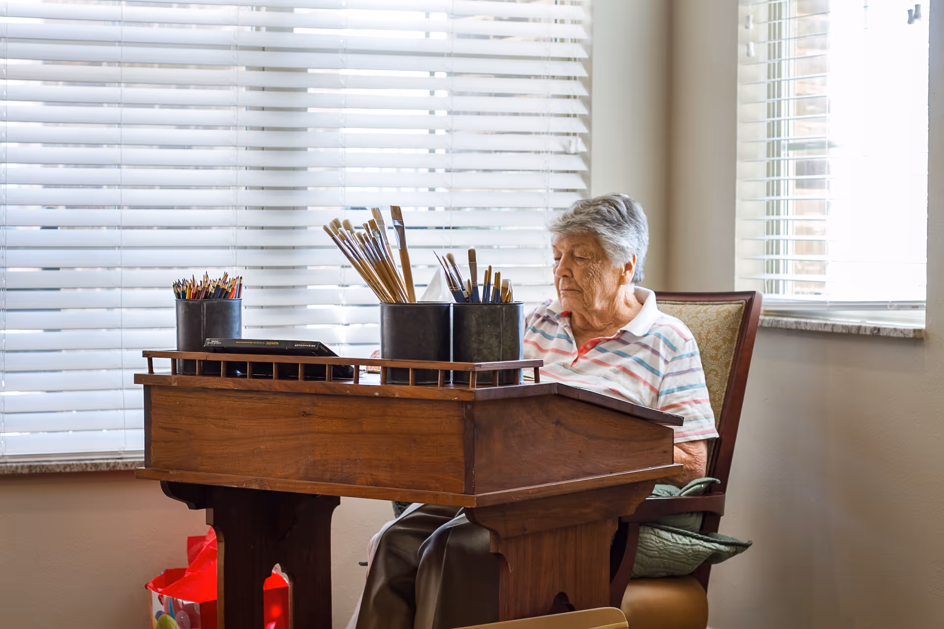 An elderly woman sitting at a wooden table with containers holding paintbrushes and colored pencils, in a room with large windows covered by white blinds.