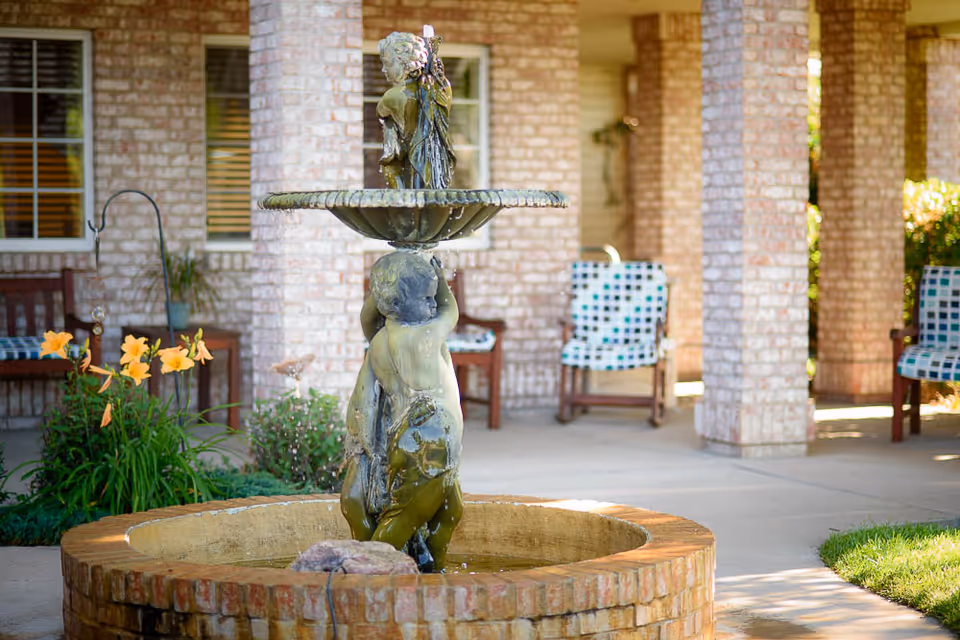 Ornamental two-tiered cherub fountain in a circular brick basin on a covered patio with chairs, columns, and flowers.