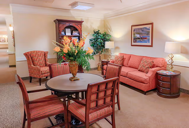 A cozy senior living common area with a round table surrounded by four wooden chairs with red striped cushions. A large vase with orange and yellow flowers is on the table. To the right, there is a red sofa with patterned pillows, two round wooden side tables with lamps, and a framed painting on the wall. In the background, there is a wooden cabinet and a green potted plant.