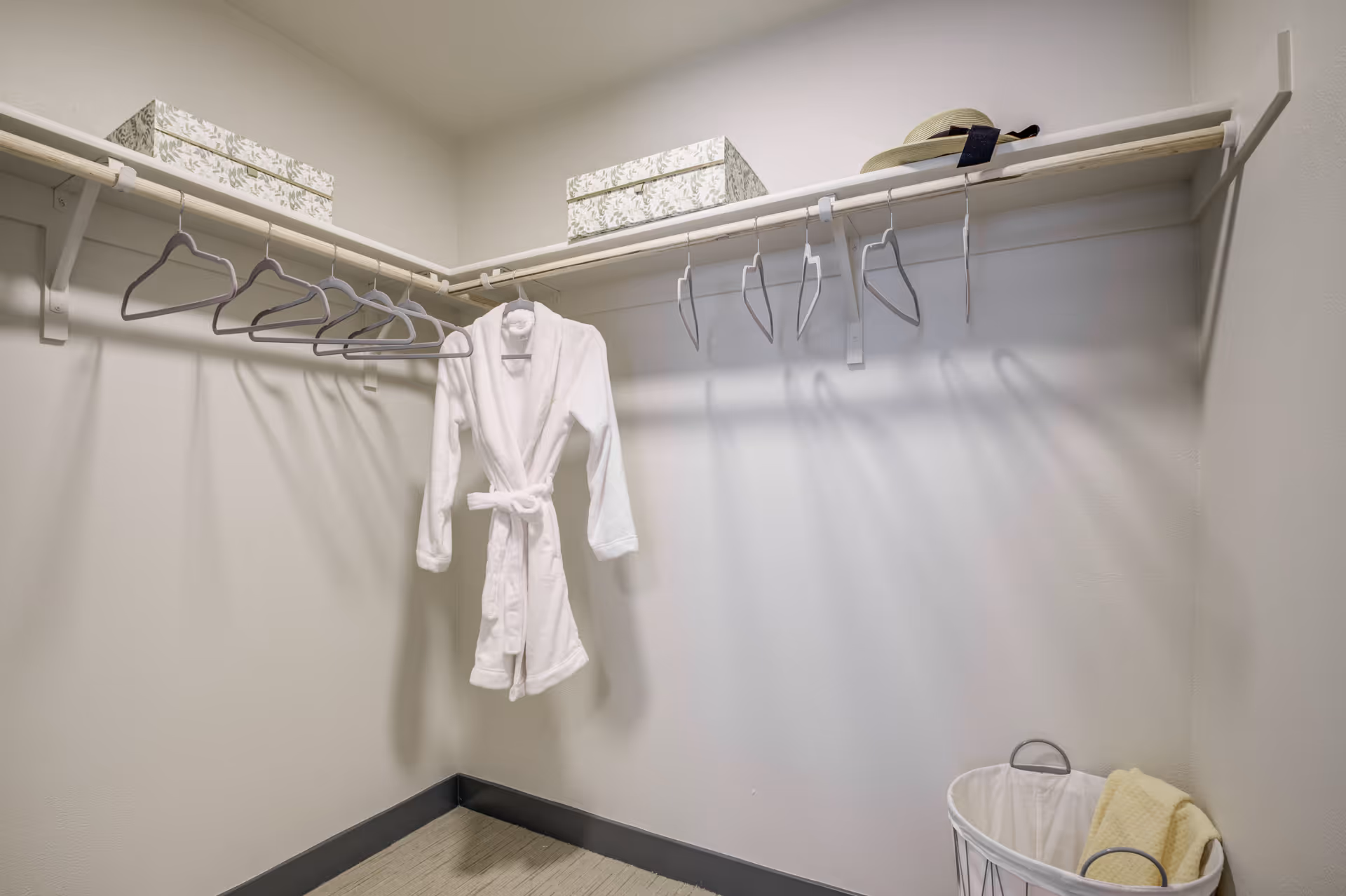 A walk-in closet with a white bathrobe hanging on a hanger, several empty hangers on a wooden rod, two decorative storage boxes on the top shelf, and a laundry basket with a yellow towel inside on the floor.
