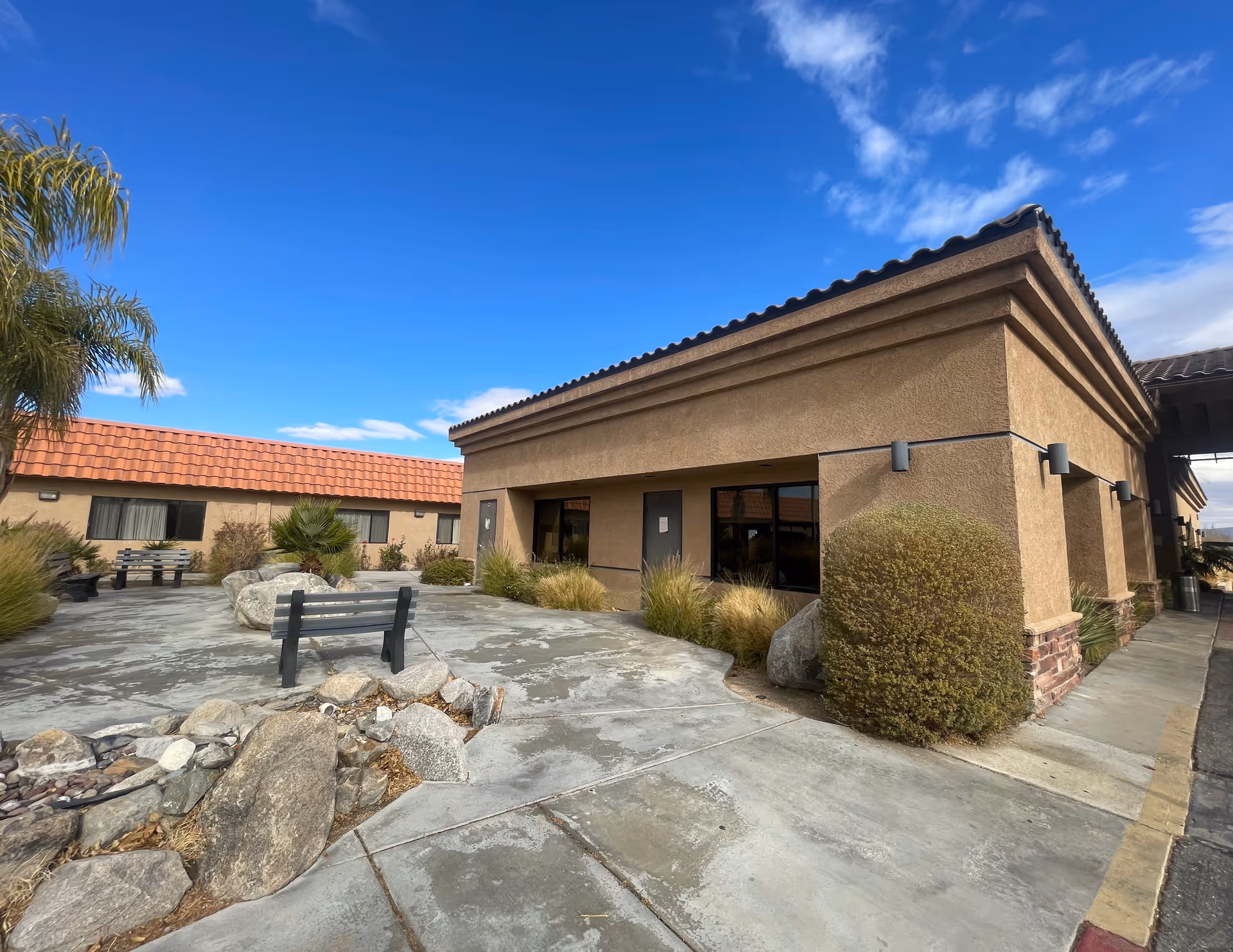 Outdoor courtyard area of Knolls West Assisted Living featuring a paved walkway, benches, rocks, shrubs, and a building with tan stucco walls and a red tile roof under a clear blue sky.