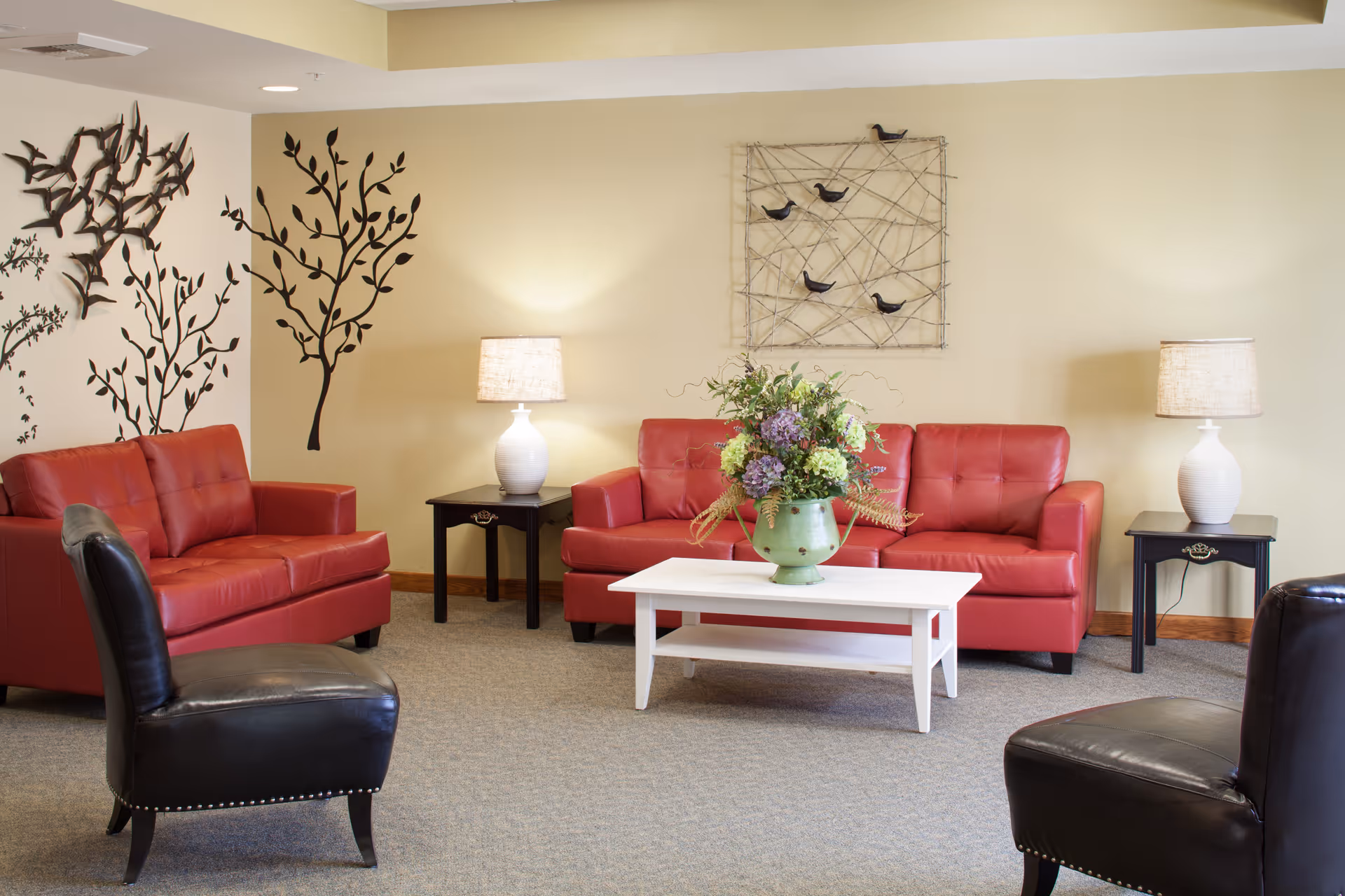 A cozy living room with two red leather sofas and two black leather chairs arranged around a white coffee table with a green vase of flowers. The walls are decorated with metal art pieces of birds and tree branches. Two side tables with white lamps flank the sofas.