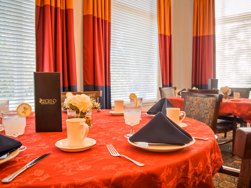 Dining table set with red tablecloth, black folded napkins on white plates, cups, glasses with lemon slices, silverware, and a small flower arrangement in a senior living facility dining room with large windows and red and orange curtains.