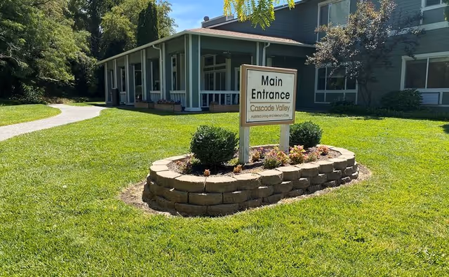 Exterior view of Cascade Valley Assisted Living and Memory Care building with a green lawn and a stone-bordered flower bed in front. A sign in the flower bed reads 'Main Entrance Cascade Valley Assisted Living and Memory Care'. The building has large windows and a covered entrance area.