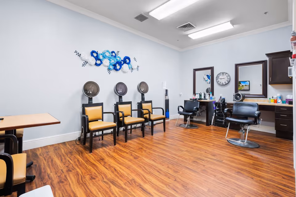 Interior view of a salon room with three hair drying chairs lined up against a wall, two styling chairs in front of mirrors, a clock on the wall, and various hair care products on the counters. The room has wood flooring and light blue walls with a decorative blue and white wall hanging.