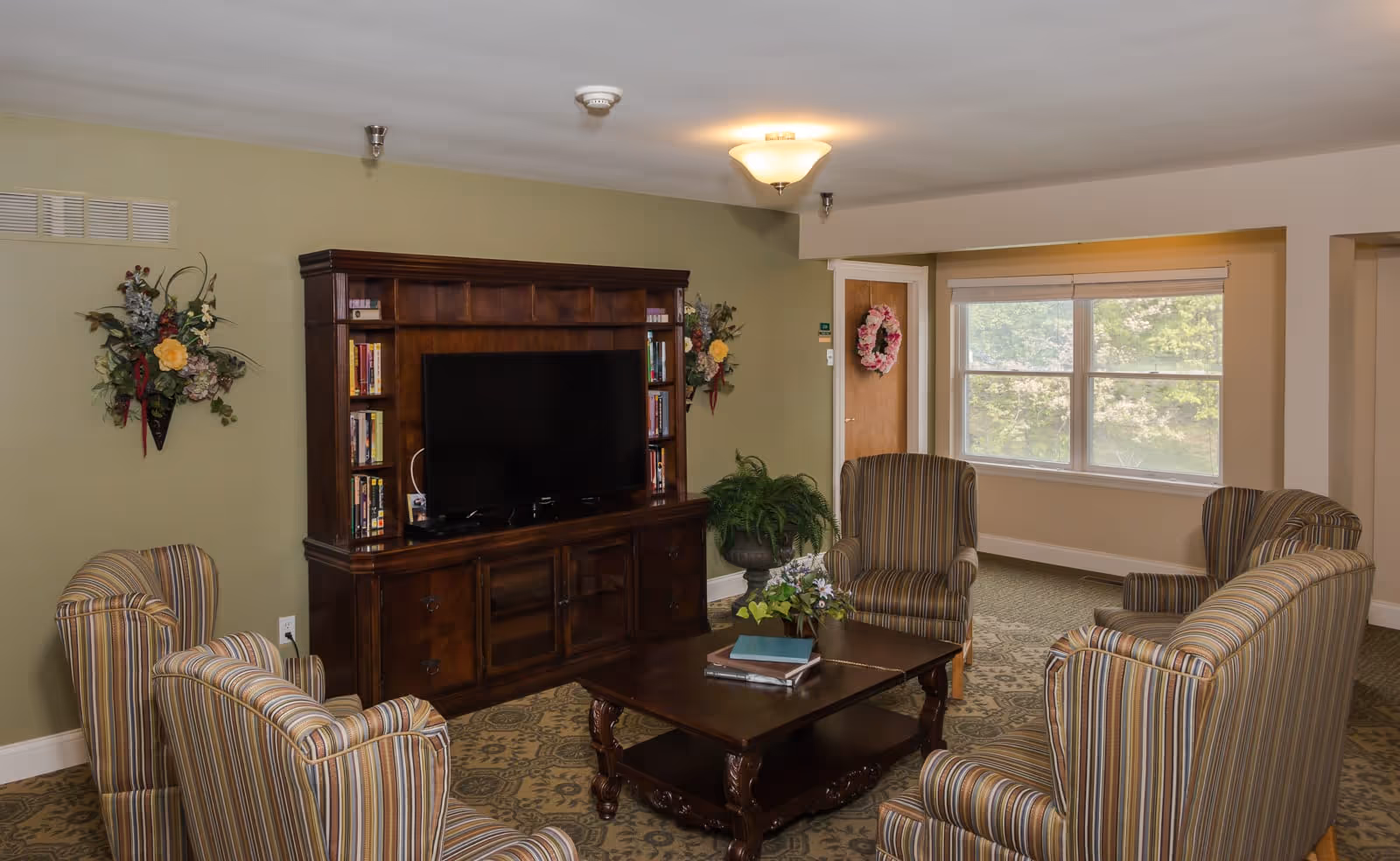 A cozy living room with six striped armchairs arranged around a dark wooden coffee table with books and a small plant. A large wooden entertainment center with a flat-screen TV and bookshelves is against a green wall. There are floral wall decorations, a window with a view of greenery, and a door with a pink floral wreath.