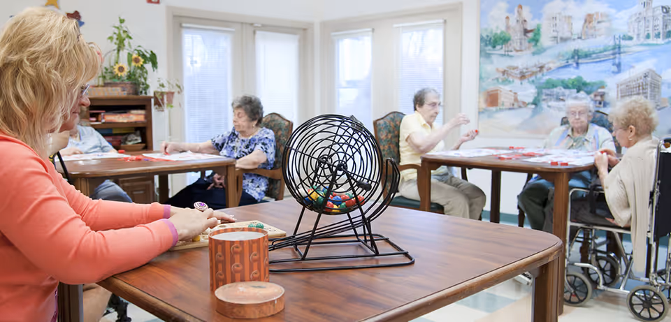 A group of elderly women sitting at tables in a bright room playing bingo. One woman in the foreground is holding bingo chips near a bingo cage filled with colorful balls. The room has large windows and a mural on the wall.