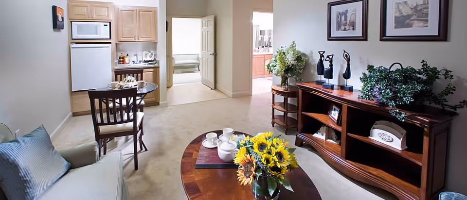 A cozy living area in The Parkview facility featuring a small round dining table with two chairs, a kitchenette with a microwave and refrigerator, a wooden coffee table with a vase of sunflowers and tea set, a comfortable armchair with a blue cushion, and a wooden sideboard decorated with plants and sculptures. The room opens into a hallway leading to other rooms.
