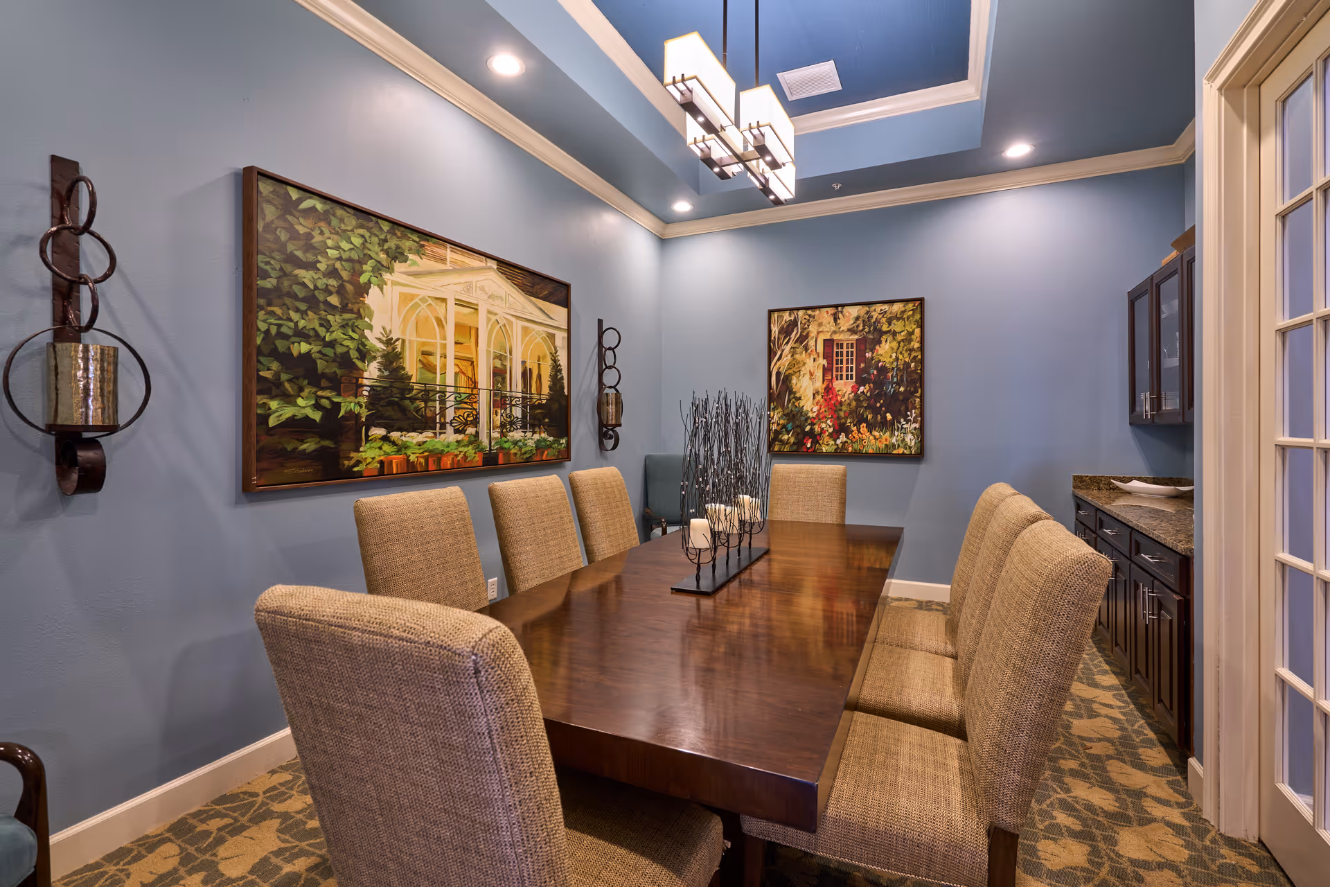 A dining room with a long wooden table surrounded by eight upholstered chairs. The walls are painted blue and decorated with two large framed paintings depicting garden scenes. There is a modern rectangular chandelier hanging from the ceiling, and a built-in cabinet with granite countertop on the right side. The floor has a patterned carpet.