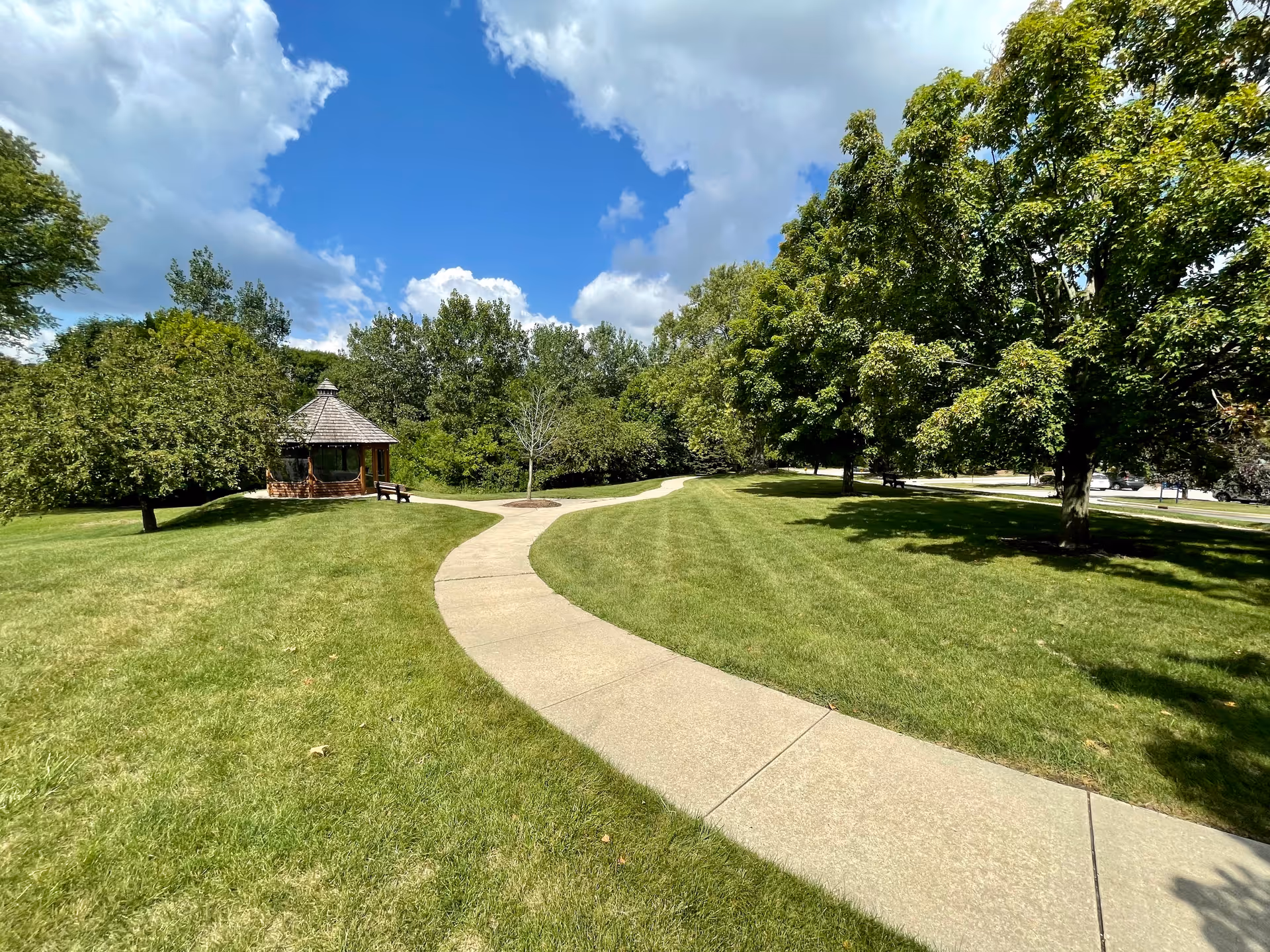 A winding concrete pathway through a well-maintained grassy area with large green trees and a small wooden gazebo with a shingled roof under a partly cloudy blue sky.