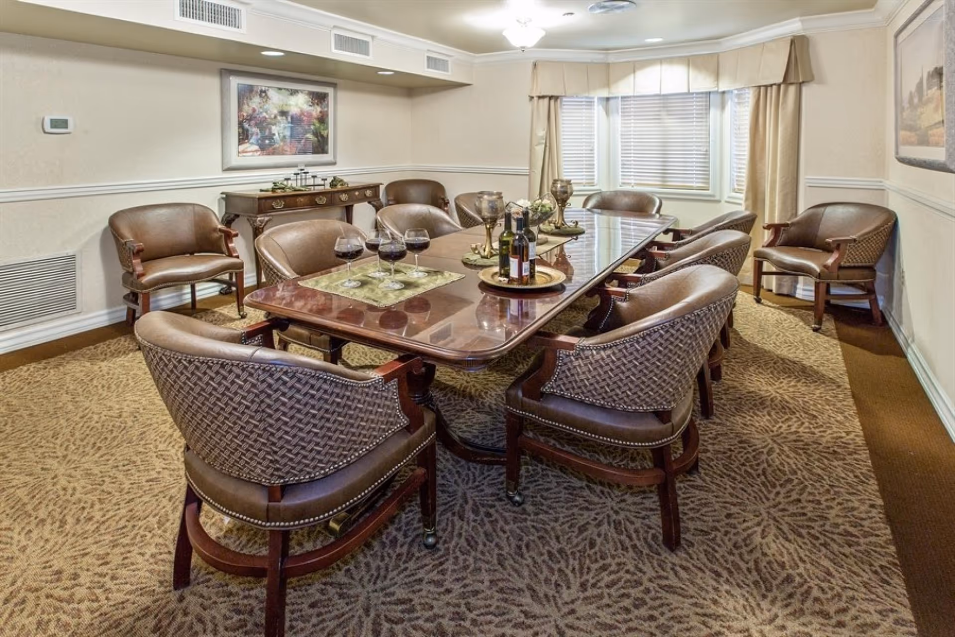 A formal dining room with a large polished wooden table surrounded by ten cushioned chairs. The table is set with four glasses of red wine, two bottles of wine, and decorative candle holders. The room has beige walls, patterned carpet, a sideboard with decorative items, and a window with blinds and curtains.