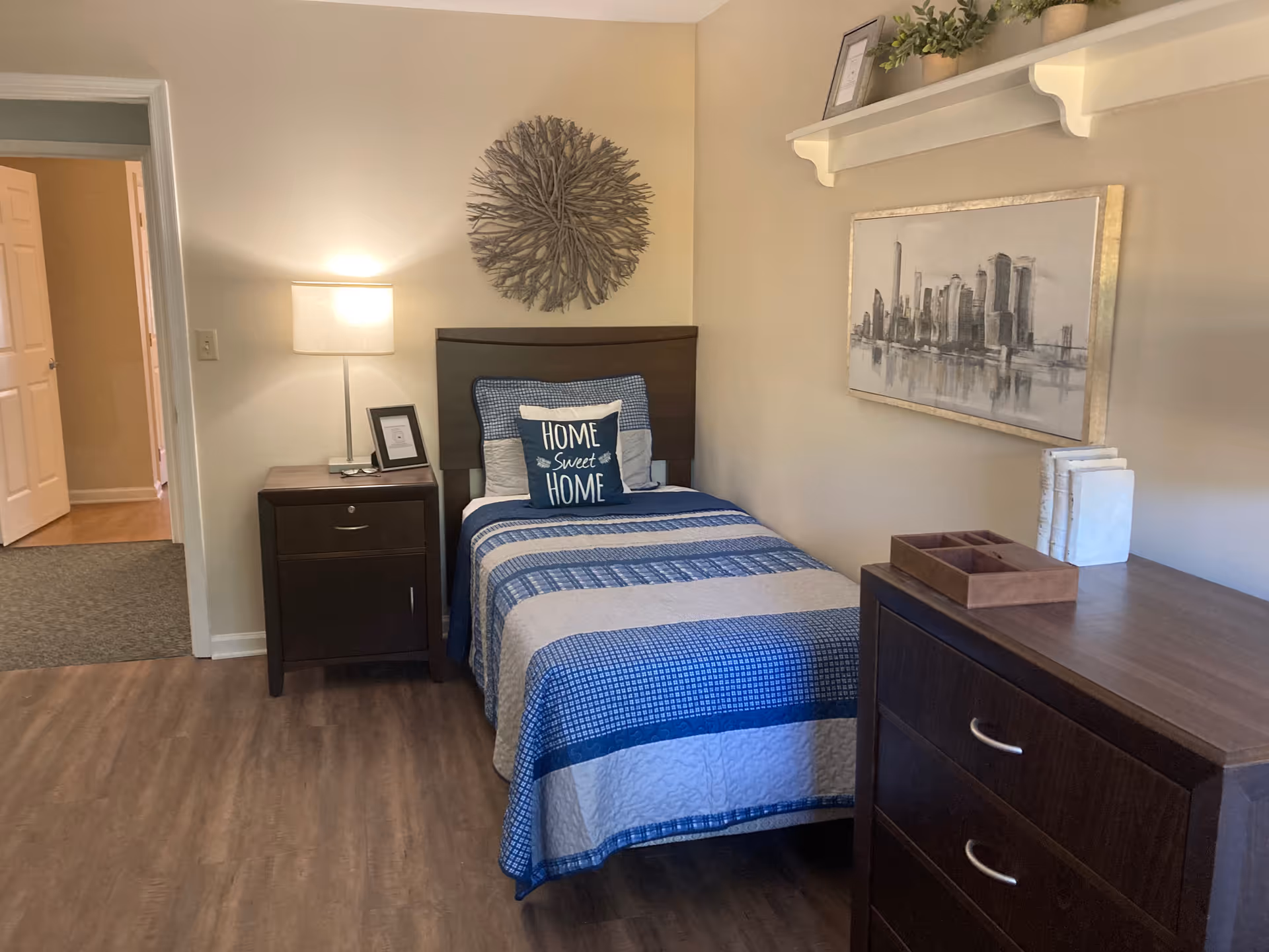 A small bedroom with a single bed covered in a blue and gray striped quilt and a pillow that says 'Home Sweet Home.' Next to the bed is a dark wooden nightstand with a lamp and a framed picture. On the right side, there is a dark wooden dresser with a tray and some books on top. Above the dresser is a framed cityscape painting and a white shelf with plants and a picture frame. The room has wood flooring and light-colored walls.