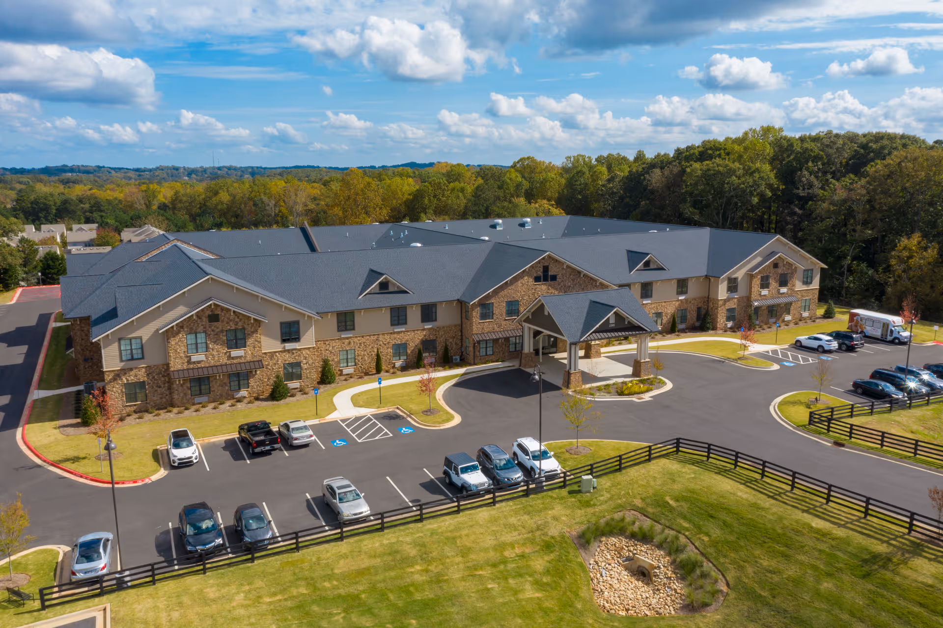 Aerial view of a large two-story senior living facility building with a stone and beige exterior, surrounded by a parking lot with several cars and a landscaped grassy area with trees and a small rock-lined pond. The sky is partly cloudy.