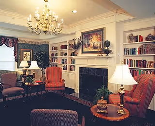 A traditional living room with upholstered armchairs, a chandelier, built-in bookshelves, and a fireplace topped by framed artwork.