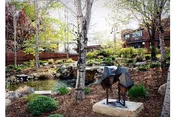 A landscaped outdoor garden area with trees, shrubs, rocks, and a small pond. There is a modern abstract metal sculpture on a stone pedestal in the foreground. In the background, a brick building is partially visible through the trees.